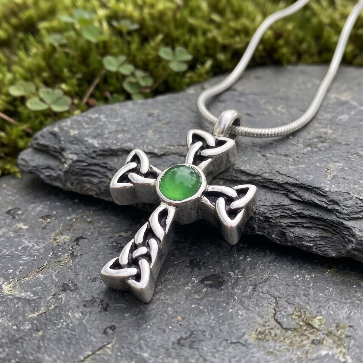 Celtic cross cremation urn necklace in silver with a green stone center, resting on dark slate beside moss and clover