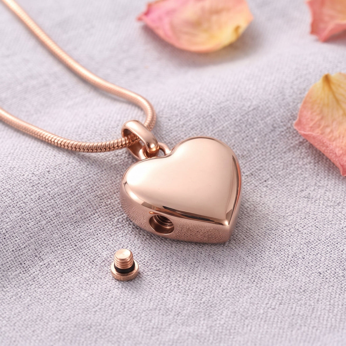Rose gold heart cremation necklace on linen with rose petals, screw removed to show the ash chamber opening