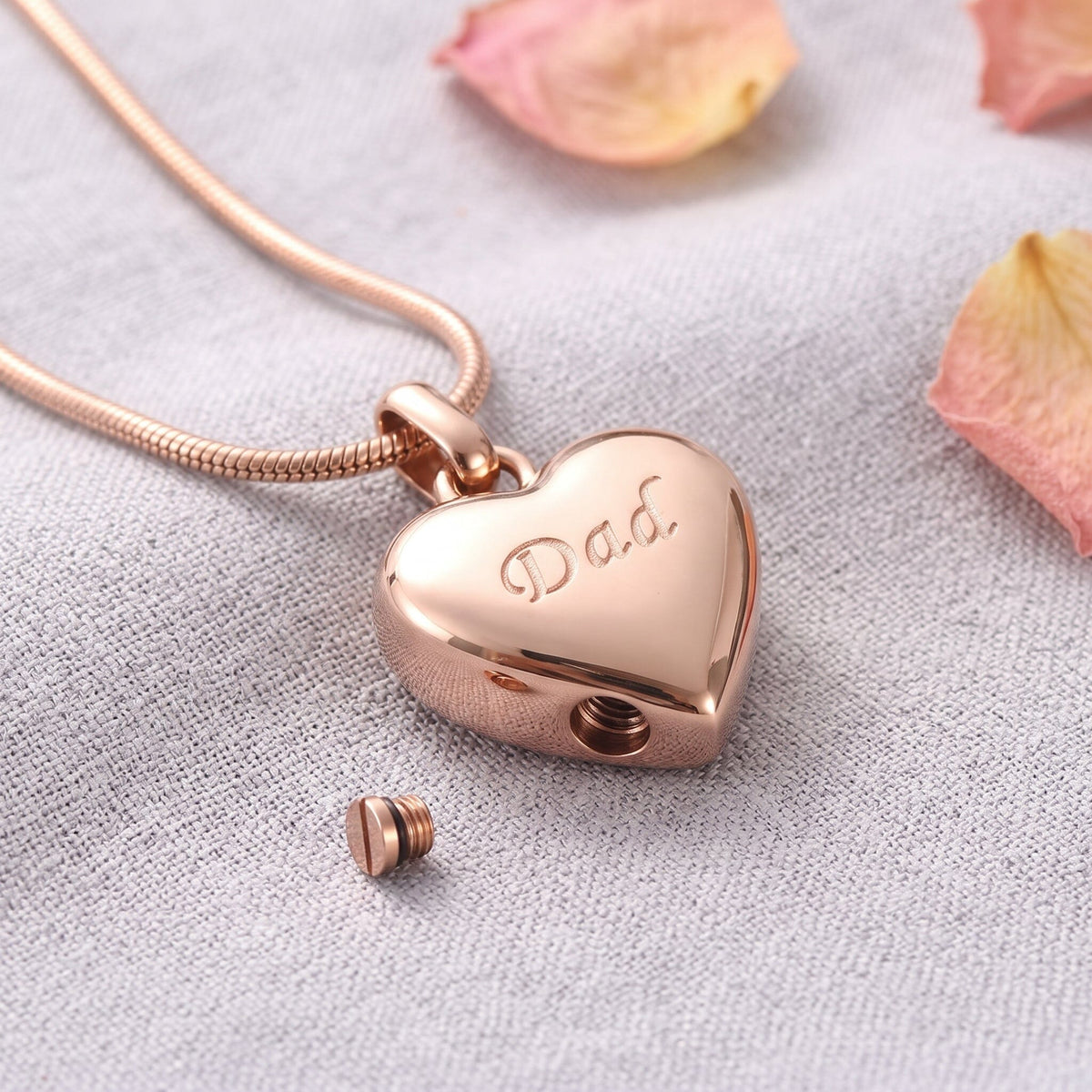 Rose gold heart cremation necklace laser engraved with Dad, resting on linen with the chamber screw beside it