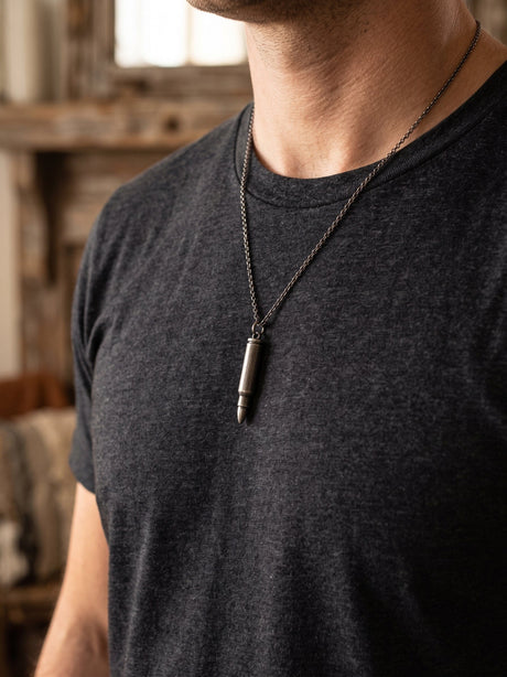 A close-up of a man wearing a dark charcoal gray t-shirt and a dark metal chain necklace with a bullet-shaped pendant, set against a blurred rustic indoor background.